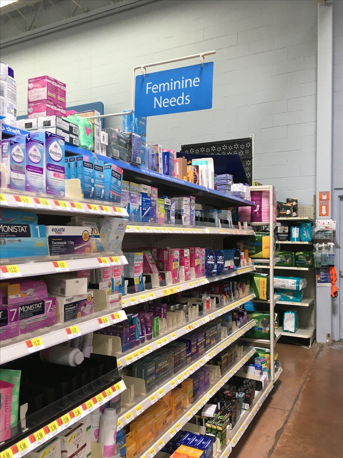 A supermarket aisle with a sign reading "Feminine Needs" above shelves of products.
